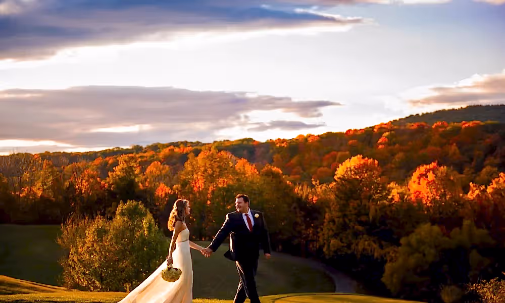 Bride and groom walking hand in hand with fall foliage behind them.