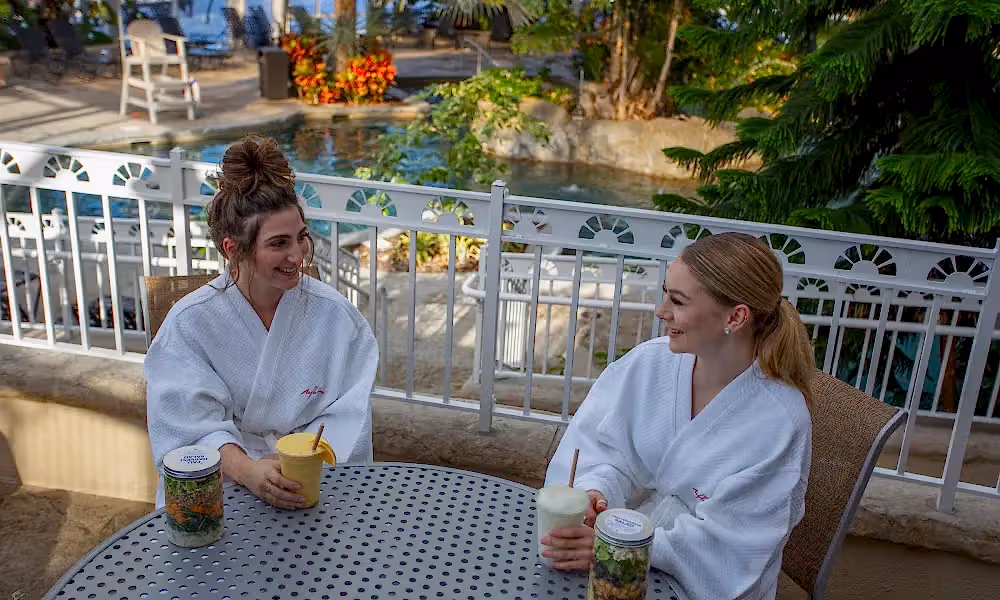 Two women sitting in white robes at a table in the Biosphere eating lunch and drinking cocktails.