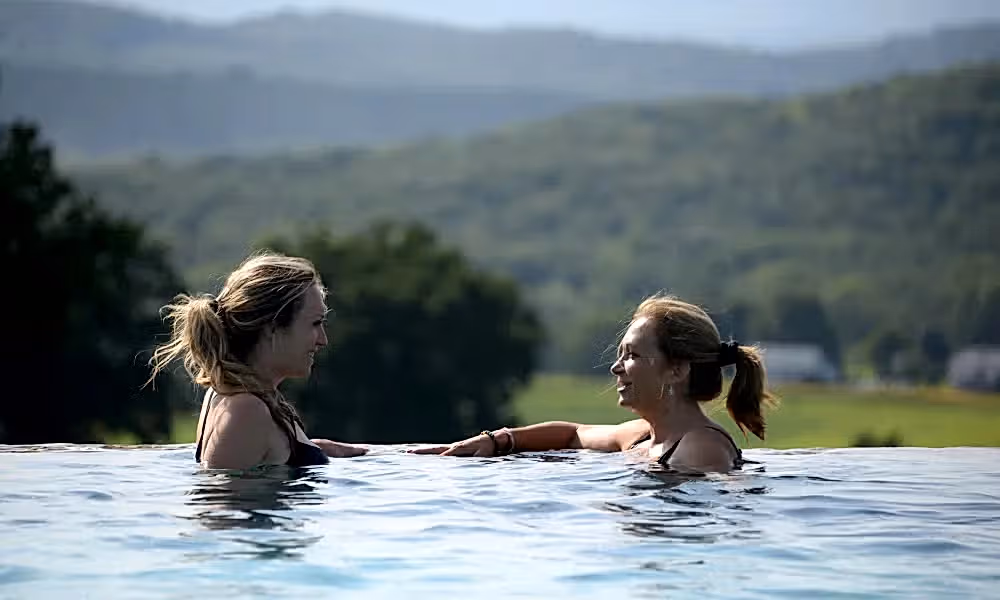 Two women speaking to one another as they relax in the vista 180 pool with mountains in background.